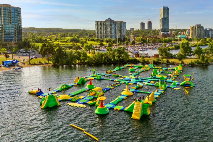 Aerial view of a colorful inflatable water park in a lake, with cityscape in the background.