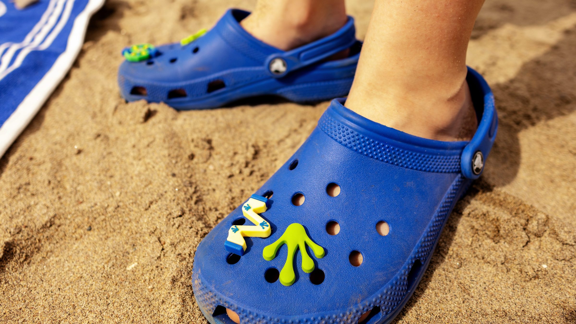 Person wearing blue clogs with frog designs on sandy beach.