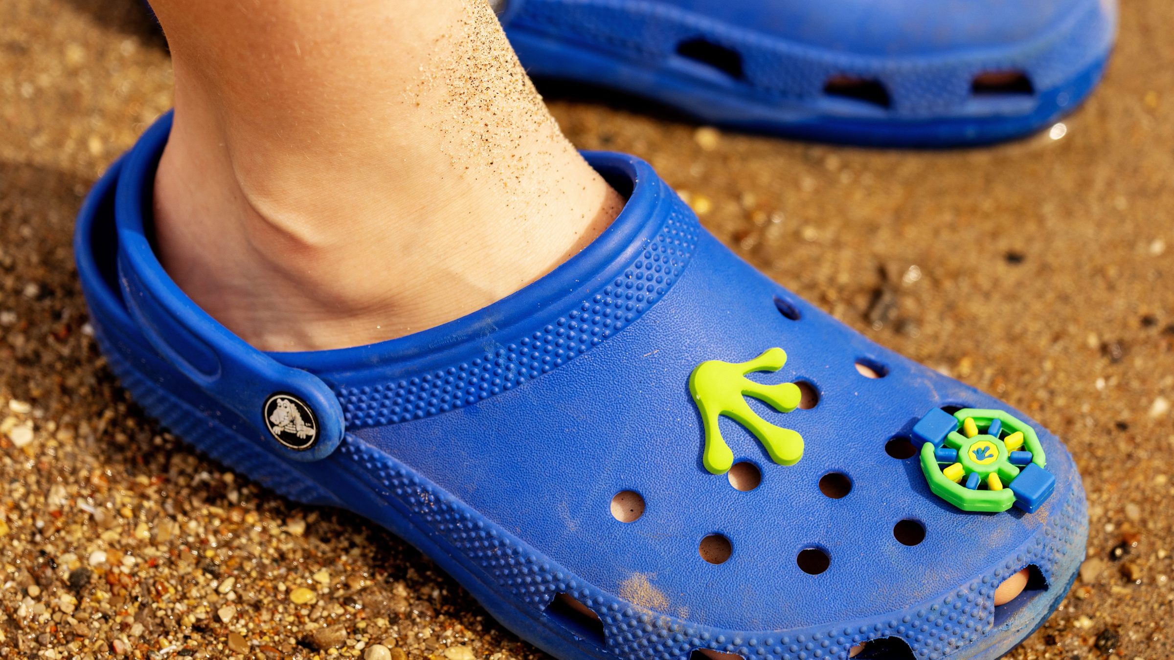Person wearing blue clogs with beach decorations standing on sand.