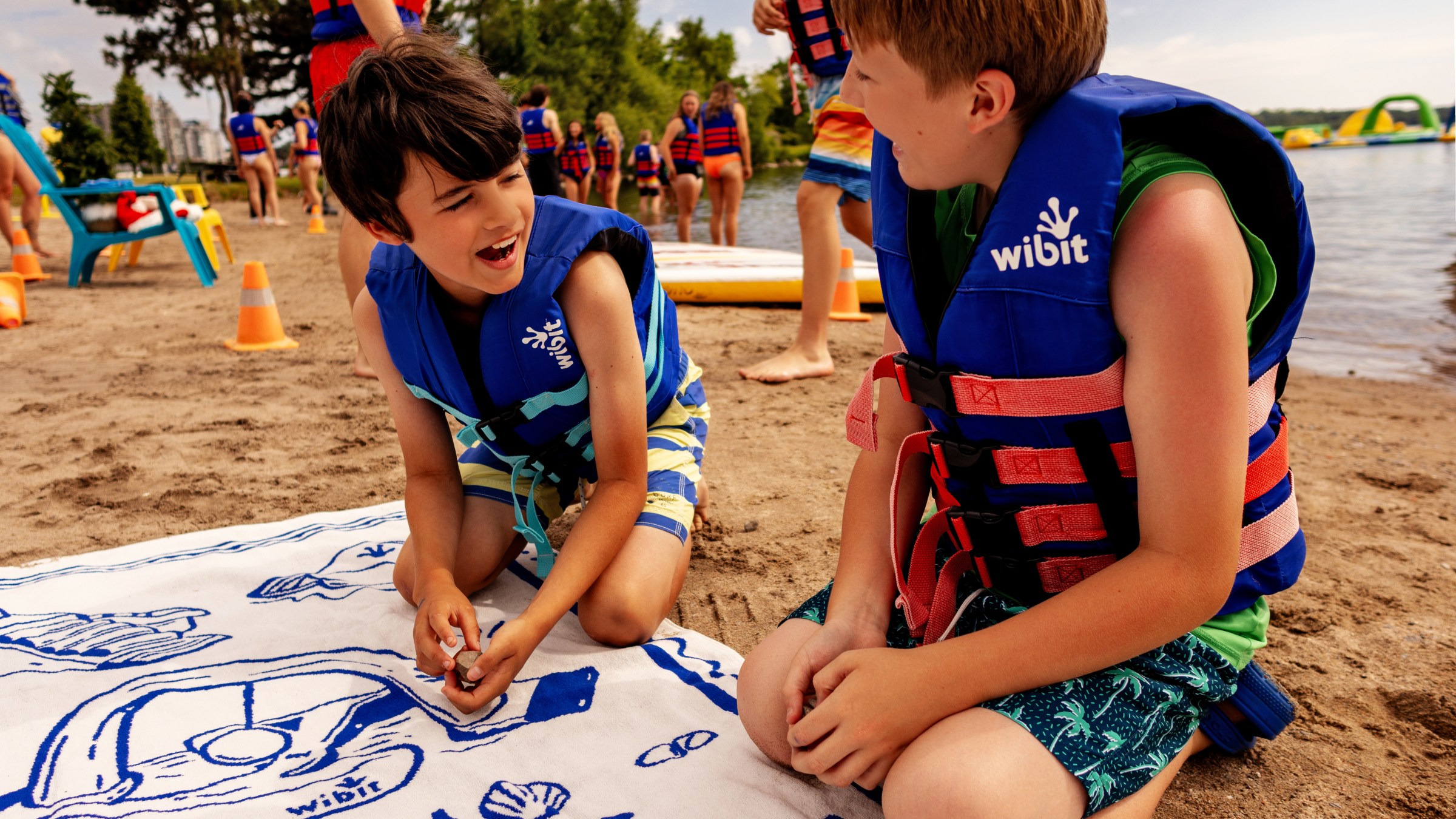 Two kids in life vests playing tic-tac-toe on beach towel by a lake.
