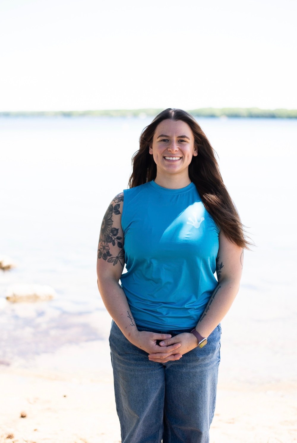 Person in blue sleeveless top and jeans stands smiling by a calm lake.